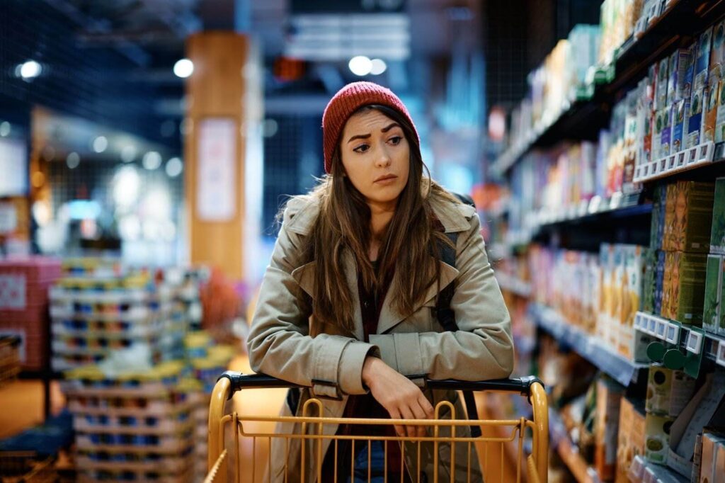 A young woman in a red beanie and tan coat leaning on a shopping cart in a grocery store.