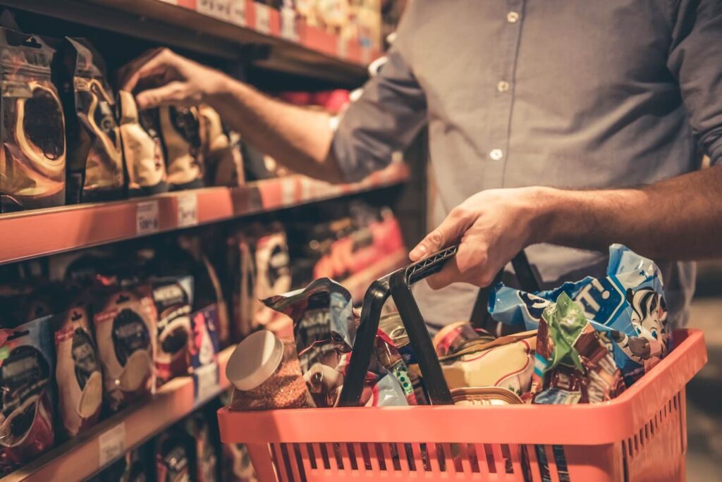 A person reaching for a packaged item on a grocery store shelf, with a red shopping basket full of items.