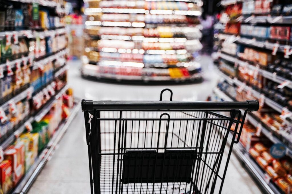 An empty black shopping cart in a grocery store aisle with stocked shelves.