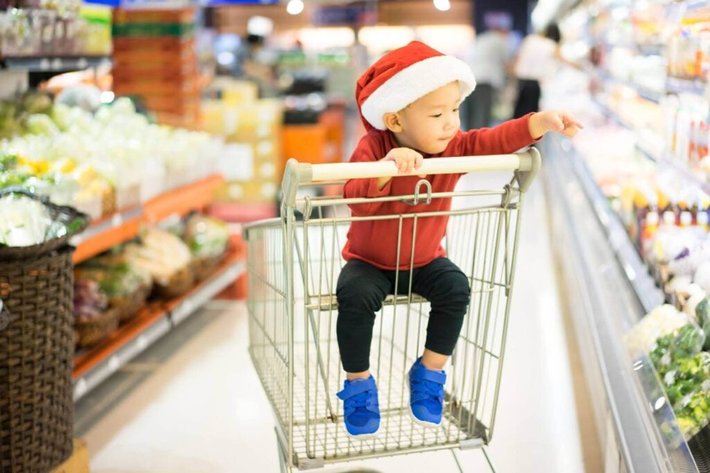 A child in a Santa hat pointing at produce from a shopping cart in a grocery store.