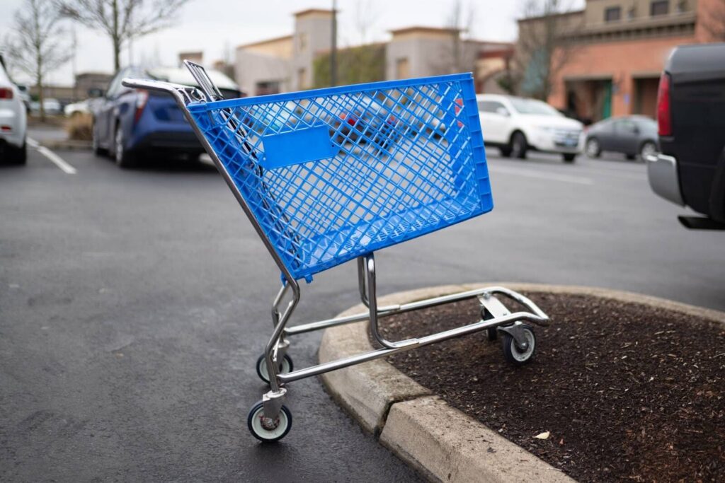 Blue shopping cart in a parking lot with cars and mulch nearby.