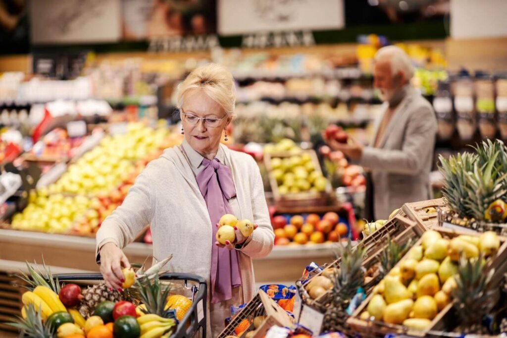An elderly woman selecting pears in a grocery store fruit section.