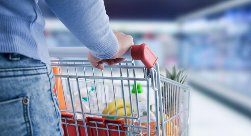 A person pushing a shopping cart filled with bananas, pineapple and packaged items through a supermarket.