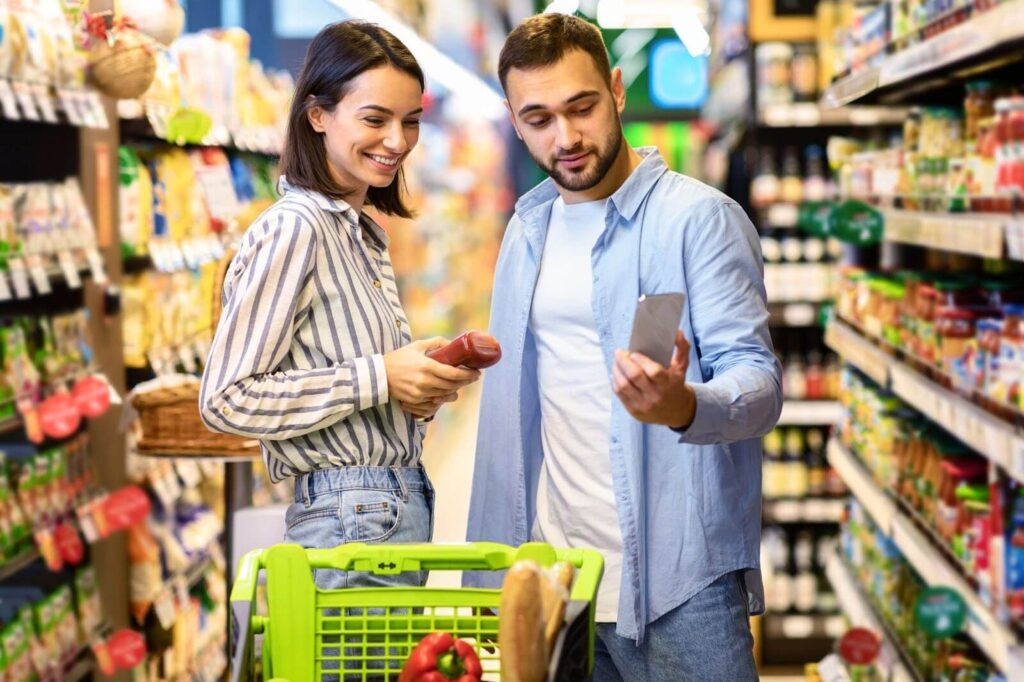 A couple in a grocery store looking at condiments and a smartphone.