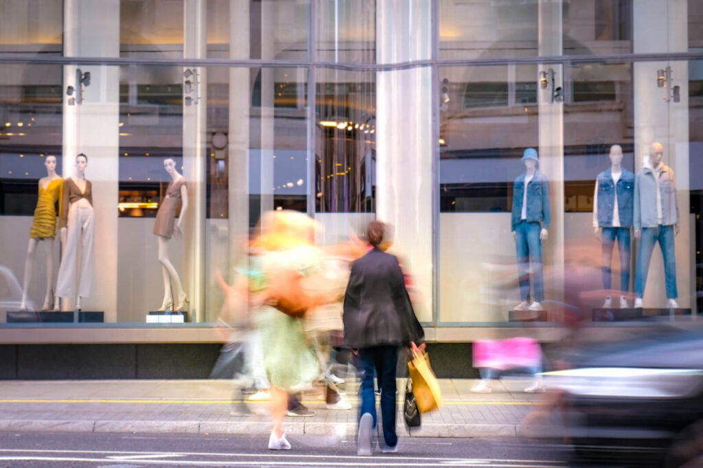 Two people standing in front of a shopping store as mannequins pose in the window display behind them.