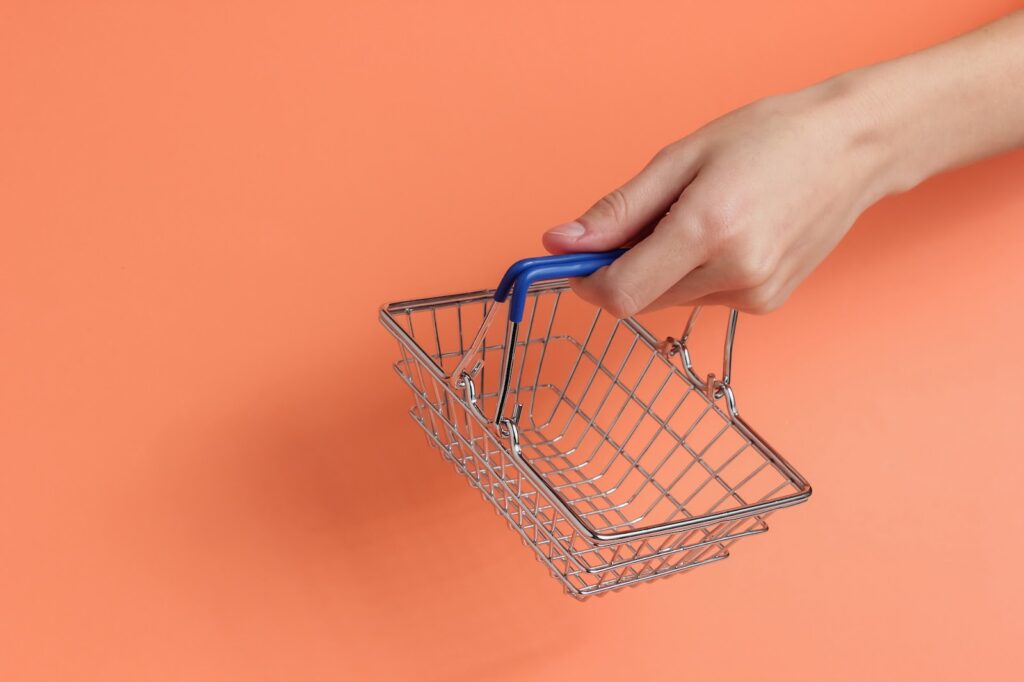 A hand holding a small metal shopping basket against a peach-colored background.