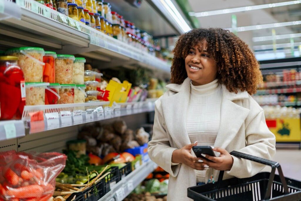 A woman with curly hair holding a shopping basket and phone in a grocery store.